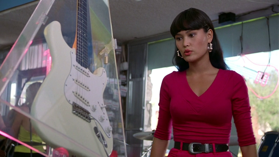 a still image from Wayne's World of actor Tia Carrere as Cassandra Wong, looking at the guitar Wayne has been coveting